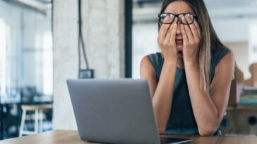 A sad adult woman wearing glasses working on a laptop.